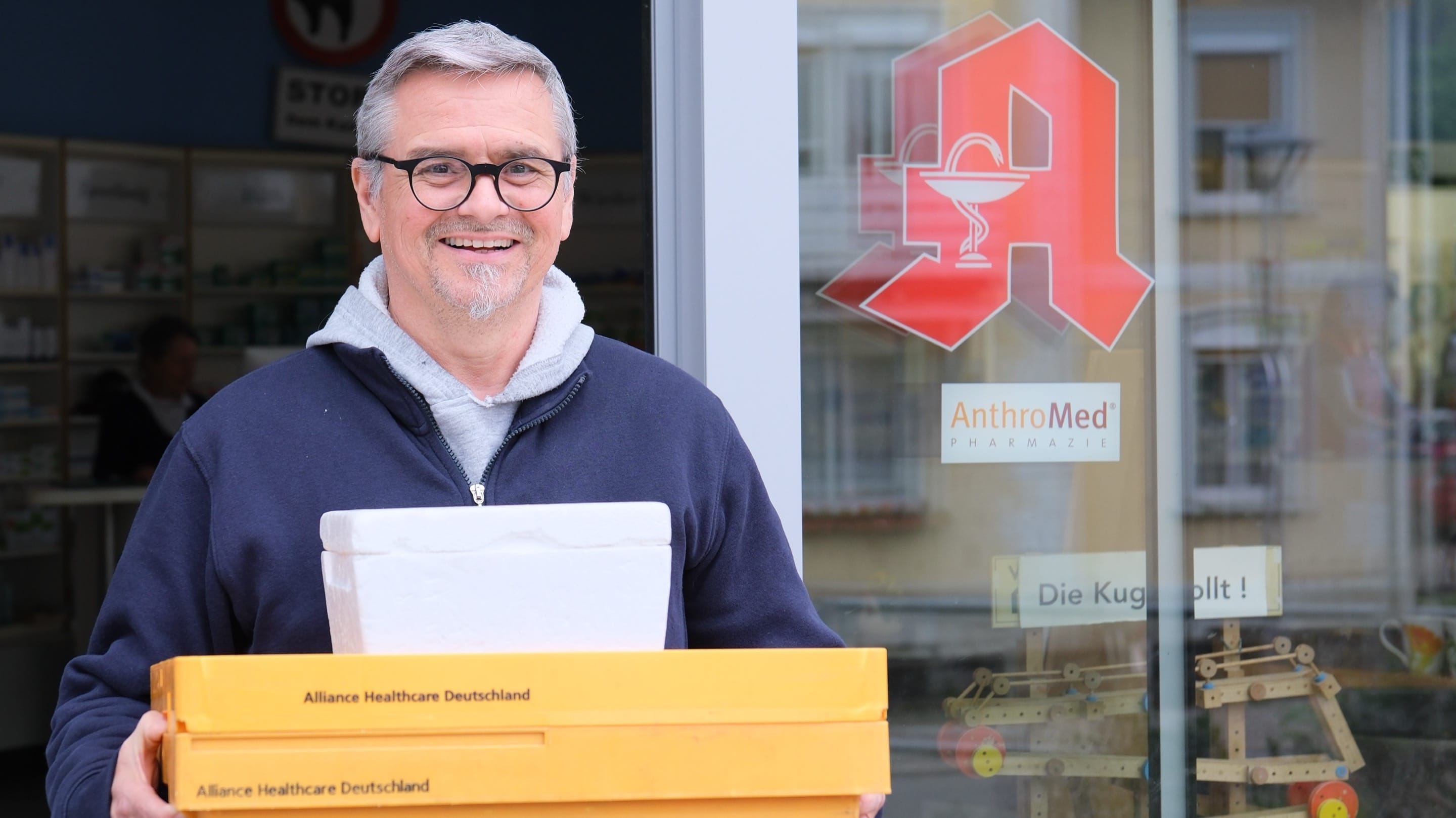 Foto unseres freundlichen Lieferanten vor der Türe der Apotheke mit zwei gelben Kisten in der Hand.